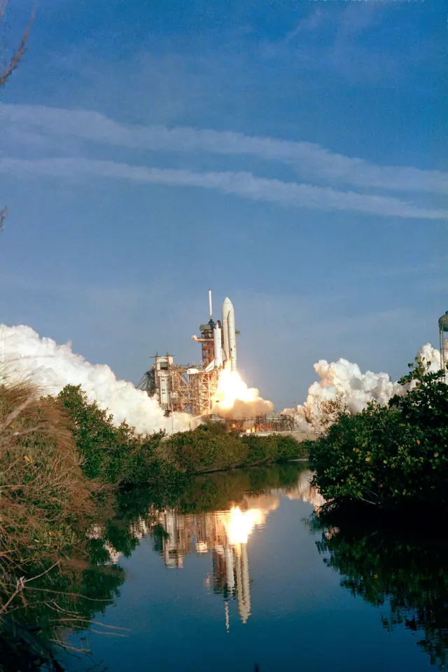 The first Space Shuttle rising from the launch pad with a white external tank, reflected in water