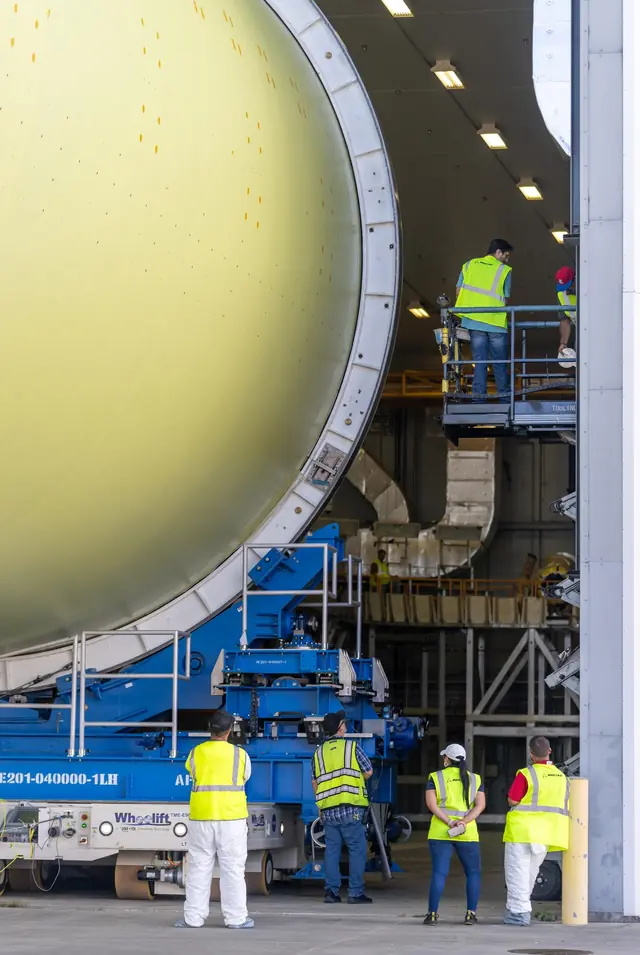 The end of a huge primed hydrogen tank being moved between work cells at Michoud, workers in safety vests beside it